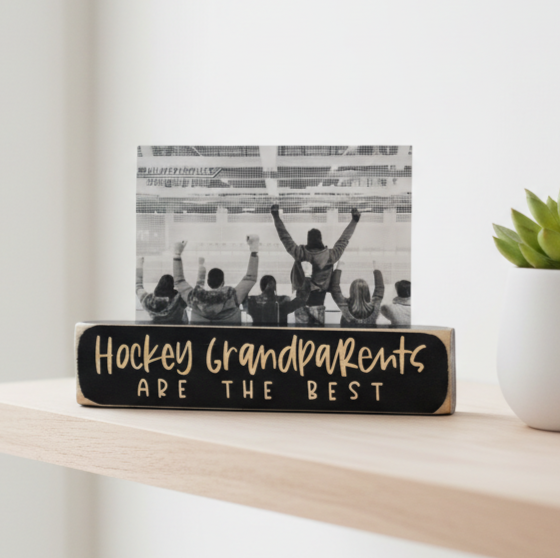 A wooden hockey-themed block sign on a light wood shelf, featuring a black base with engraved lettering that reads 'Hockey Grandparents ARE THE BEST' and a black-and-white photo of people cheering in the background.