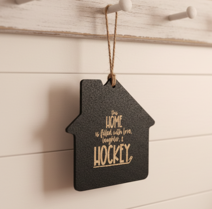 A house-shaped black hanging sign with tan lettering that reads 'The home is filled with love, laughter, & HOCKEY,' hanging from a rustic white peg on a white paneled wall.