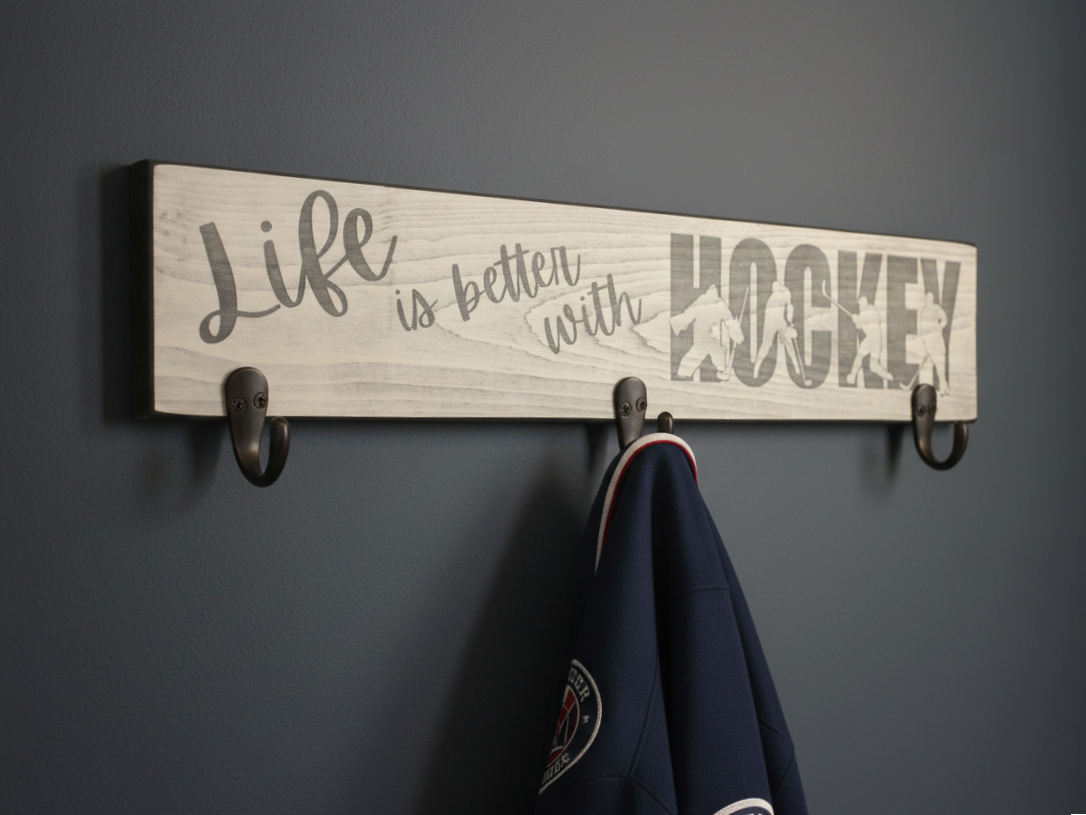 A rustic white-painted wooden wall sign with three dark metal hooks, featuring the phrase 'Life is better with HOCKEY' in gray script and block lettering. A navy blue hockey jersey is hanging from one of the hooks against a dark gray wall.