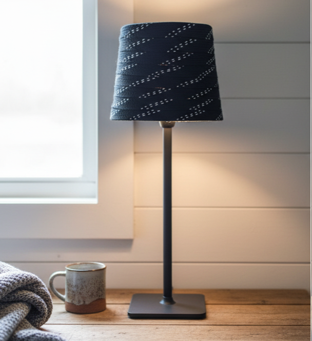 A black table lamp with black hockey laces shade  set on a rustic table next to a ceramic mug in a cozy room setting.