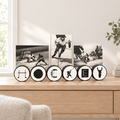 A set of  connected hockey pucks, spelling out  "HOCKEY" with unique hockey letters.   This displays 3 hockey photos and is sitting on a natural wood table with plants and books in a cozy room with light pouring in.