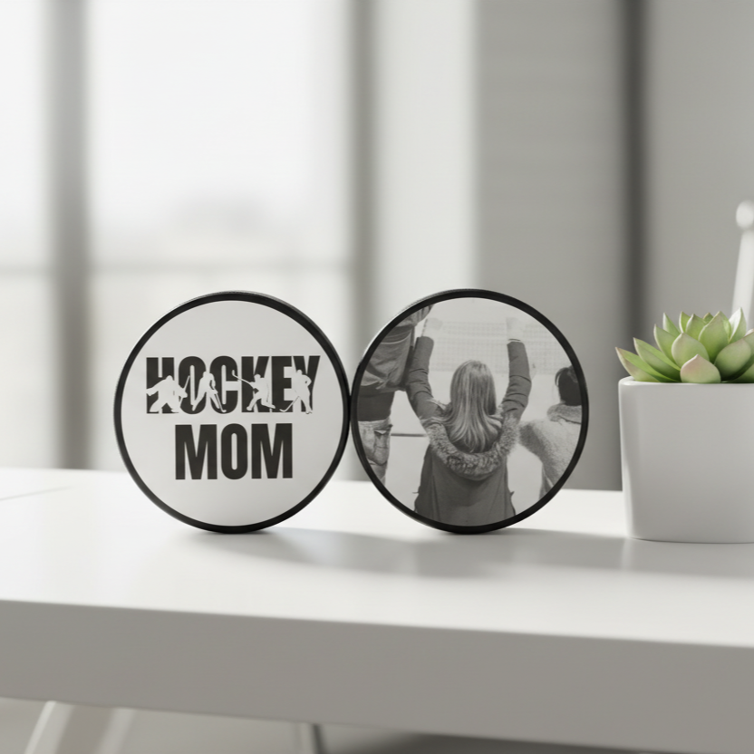 Two connected photo hockey pucks on a white table. The left sign has a white background with black text reading 'HOCKEY MOM' and hockey player silhouettes.  The other puck has a custom photo on it.