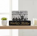 A rectangular black wooden sign with engraved lettering reading 'Hockey Mom' sits on a light wood shelf, holding a black-and-white photo of hockey players with raised arms. A small potted succulent is on the left and a white candle in a glass holder is on the right, against a bright indoor background.