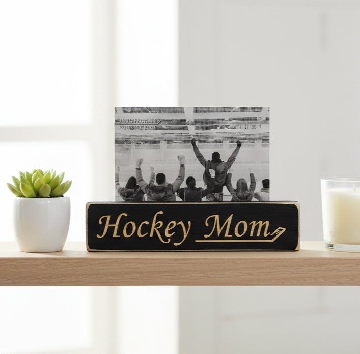 A rectangular black wooden sign with engraved lettering reading 'Hockey Mom' sits on a light wood shelf, holding a black-and-white photo of hockey players with raised arms. A small potted succulent is on the left and a white candle in a glass holder is on the right, against a bright indoor background.