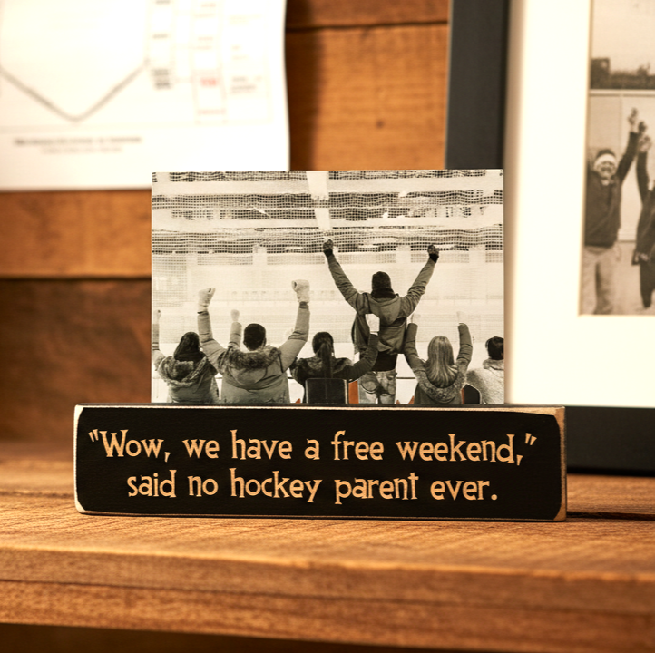 A rectangular black wooden plaque with engraved lettering sits on a wooden surface. The plaque reads, "Wow, we have a free weekend, said no hockey parent ever.