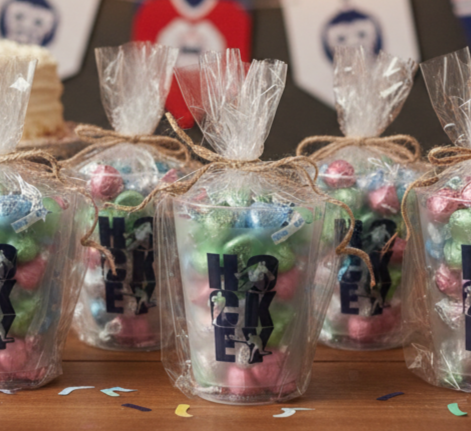 Several clear plastic cups filled with wrapped candies and tied with twine, printed with the word "HOCKEY" in large black letters, arranged on a wooden table at a party.