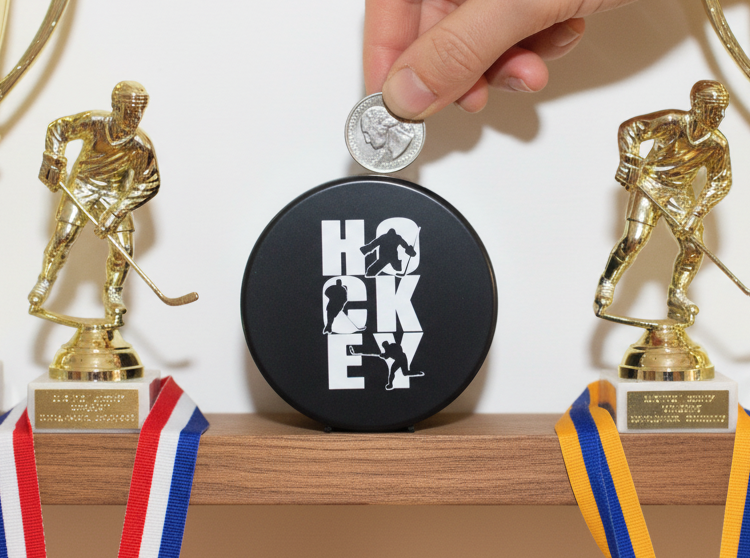 A black round hockey puck-shaped bank with white 'HOCKEY' lettering sits on a wooden shelf, with a hand placing a coin on top. Two gold hockey player trophies with red, white, and blue ribbons flank it against a plain light background.