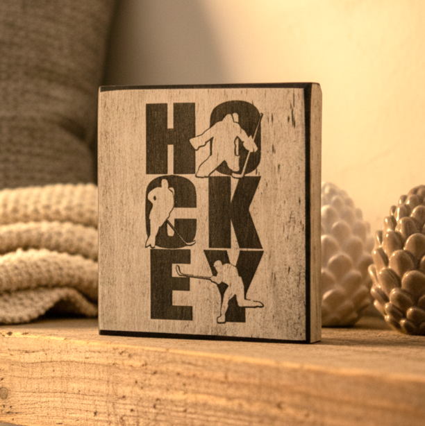 A square wooden block sign with a distressed whitewash finish and black laser-cut lettering spelling 'HOCKEY' with silhouettes of hockey players, displayed on a wooden surface with a cozy background.