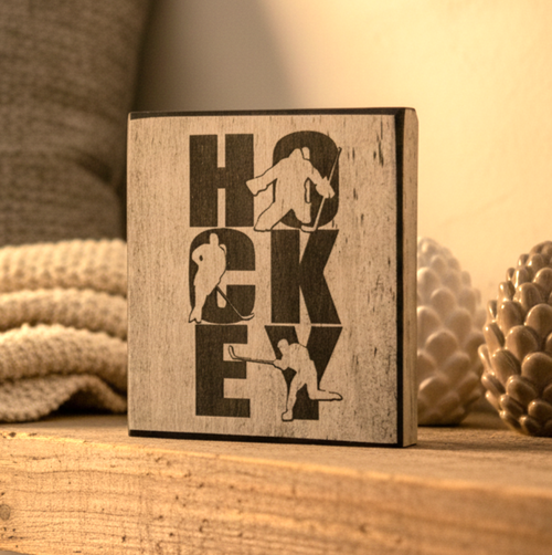A square wooden block sign with a distressed whitewash finish and black laser-cut lettering spelling 'HOCKEY' with silhouettes of hockey players, displayed on a wooden surface with a cozy background.
