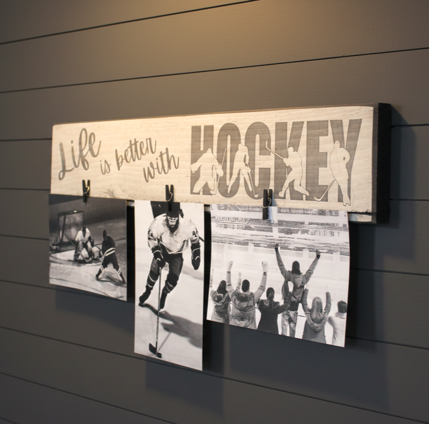 A rustic wooden hockey wall sign with the phrase 'Life is better with HOCKEY' and silhouettes of players, shown hanging on a dark gray paneled wall with three printed hockey photos clipped below.