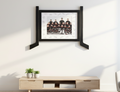 A framed sports team photograph with black decorative hockey stick side brackets, mounted on a white wall above a light wood console table with small potted plants.