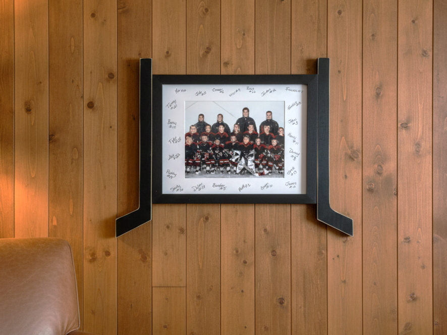 A black-framed horizontal team photograph with hockey sticks on both sides, signed by the players, mounted on a warm wood-paneled wall.