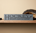 A rectangular dark gray wooden hockey sign with the words 'HOCKEY WHAT'S NOT TO LOVE?' displayed on a wooden shelf next to a brown leather glove.