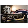 A rectangular black wooden hockey-themed sign with engraved lettering that reads 'They can dangle and shoot, but there will be NO CELLY!' and an engraved goalie mask graphic, shown in a softly blurred indoor setting with a hockey mask and flag in the background.
