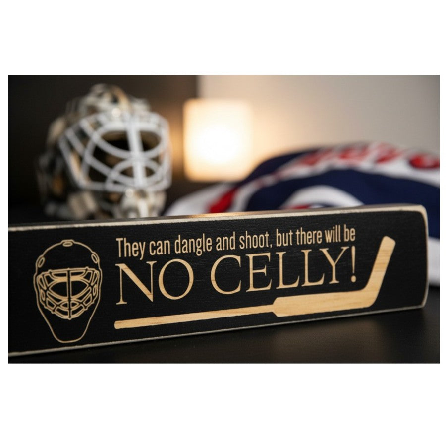 A rectangular black wooden hockey-themed sign with engraved lettering that reads 'They can dangle and shoot, but there will be NO CELLY!' and an engraved goalie mask graphic, shown in a softly blurred indoor setting with a hockey mask and flag in the background.