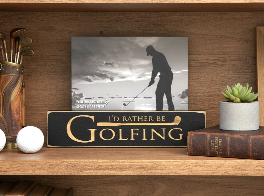 A framed-style decorative golf sign on a wooden shelf, featuring a black-and-white photo of a golfer and the text 'I'D RATHER BE GOLFING' in engraved lettering on a dark plaque. Surrounding props include a golf bag with clubs, golf balls, a small succulent, and a vintage book.