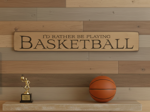 A rectangular wooden sign with the engraved text 'I'D RATHER BE PLAYING BASKETBALL' displayed on a rustic wood plank wall above a shelf with a basketball and a small trophy.