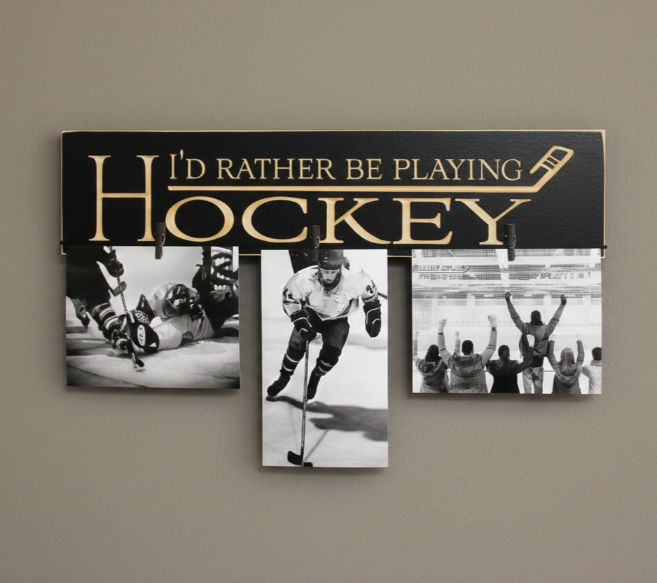 A black hockey-themed photo collage sign with engraved lettering reading 'I'D RATHER BE PLAYING HOCKEY' and three hanging black-and-white photos of hockey players and fans, mounted on a beige wall.
