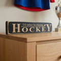A black rectangular wooden sign with engraved lettering that reads 'I'd rather be playing HOCKEY', displayed on a light wood dresser in a room setting.