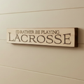 Wood beige Lacrosse sign with carved lettering which reads "I'd rather be playing LACROSSE" hung on a wall with soft lighting