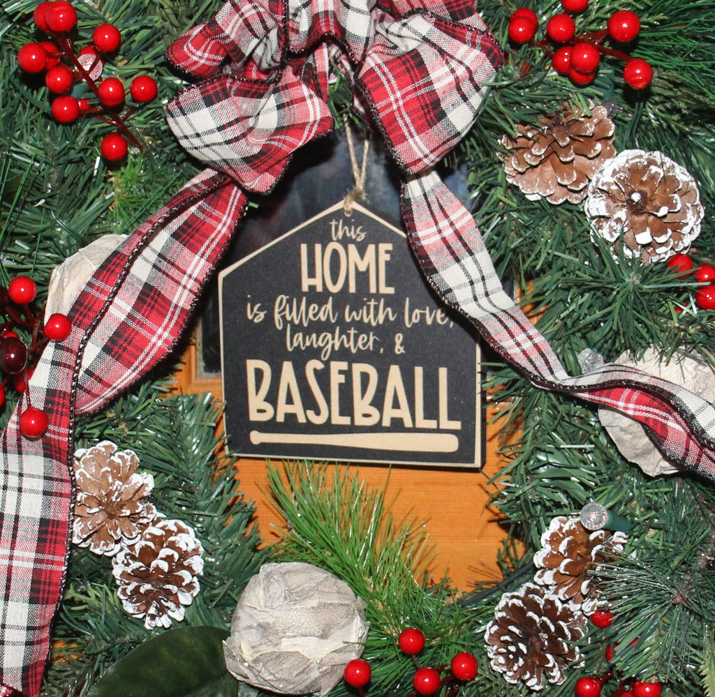 May include: A Christmas wreath features a red and black plaid ribbon, pinecones, berries, and a house-shaped ornament. The ornament reads: 'This Home is filled with love, laughter, & Baseball'. The wreath is a festive blend of traditional Christmas elements and baseball-themed decorations, perfect for a sports fan's holiday decor.