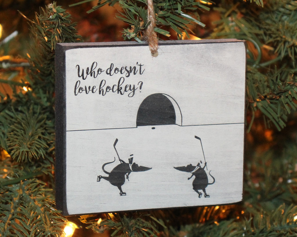 May include: A white wooden Christmas ornament with black text and a black and white illustration. The text reads 'Who doesn't love hockey?' The illustration shows two mice playing hockey on a white ice rink with a black goal net.