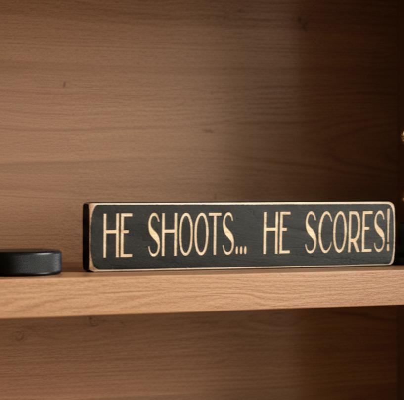 Black wooden sign with the saying "He Shoots... He Scores!" sitting on a shelf with a puck next to it