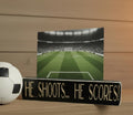 Black wood photo sign with engraved lettering reading "He Shoots... He Scores!" holding a soccer photo and sitting on a shelf along with soccer decor.