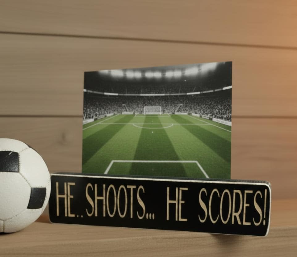 Black wood photo sign with engraved lettering reading "He Shoots... He Scores!" holding a soccer photo and sitting on a shelf along with soccer decor.