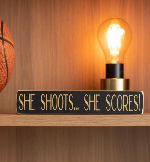 Black wood sign with engraved lettering reading "She Shoots... She Scores!", sitting on a shelf along with basketball decor.
