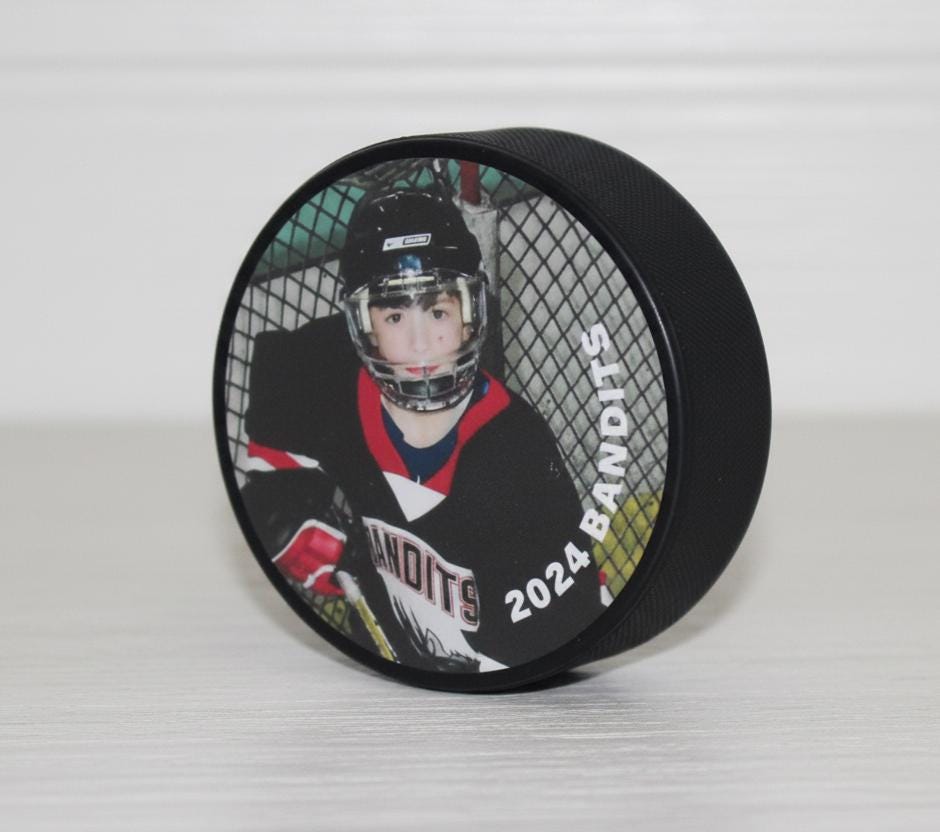 Hockey puck with a photo of a hockey player and the name of the team and year, sitting on a white table and white shiplap background.