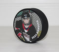 Hockey puck with a photo of a hockey player and the name of the team and year, sitting on a white table and white shiplap background.