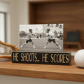 Black wood photo sign with engraved lettering reading "He Shoots... He Scores!", sitting on a shelf along with lacrosse decor.