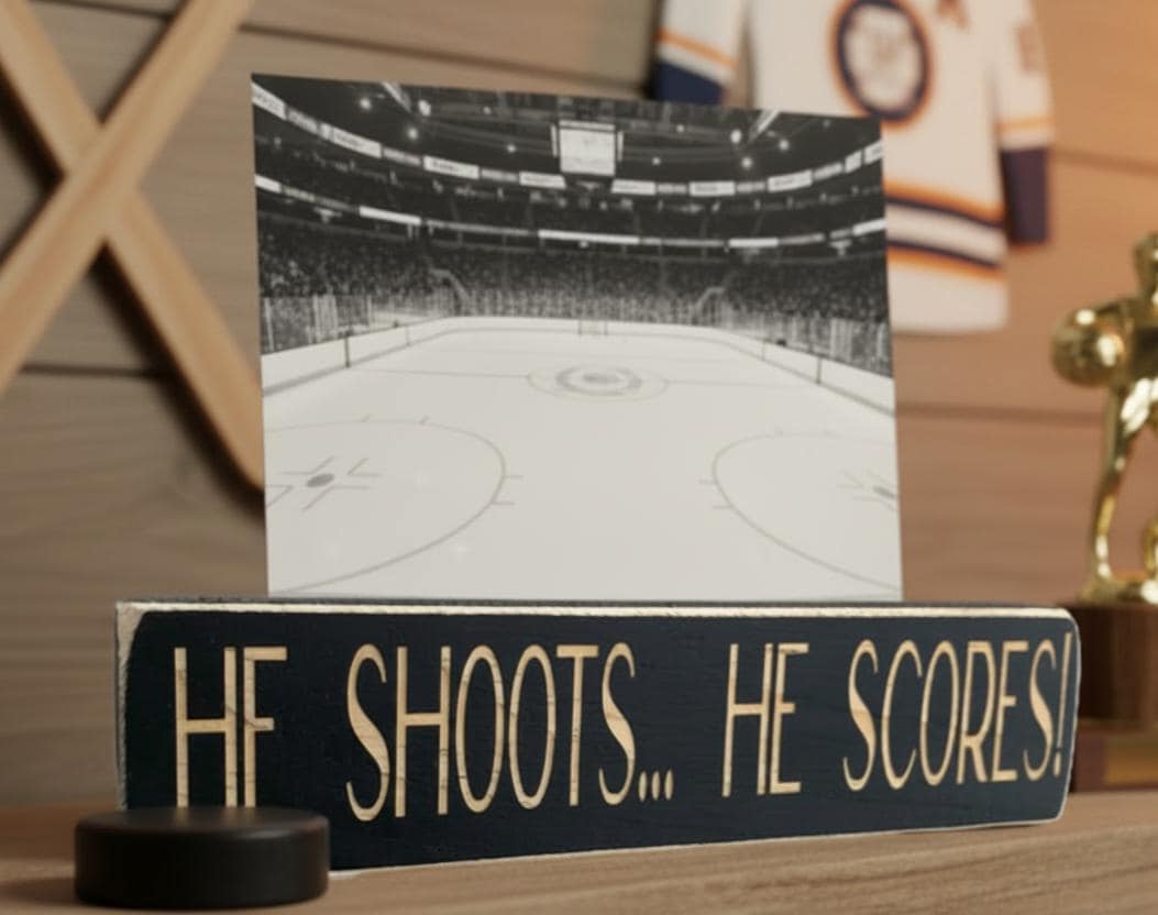 Wood photo sign with the words "He Shoots... He Scores!" engraved on the sign and it is holding a hockey photo and sitting on a shelf along with hockey decor.
