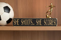 Black wood sign with engraved lettering reading "She Shoots... She Scores!", sitting on a shelf along with soccer decor.