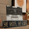 Black wood photo sign with engraved lettering reading "She Shoots... She Scores!", sitting on a shelf along with basketball decor.