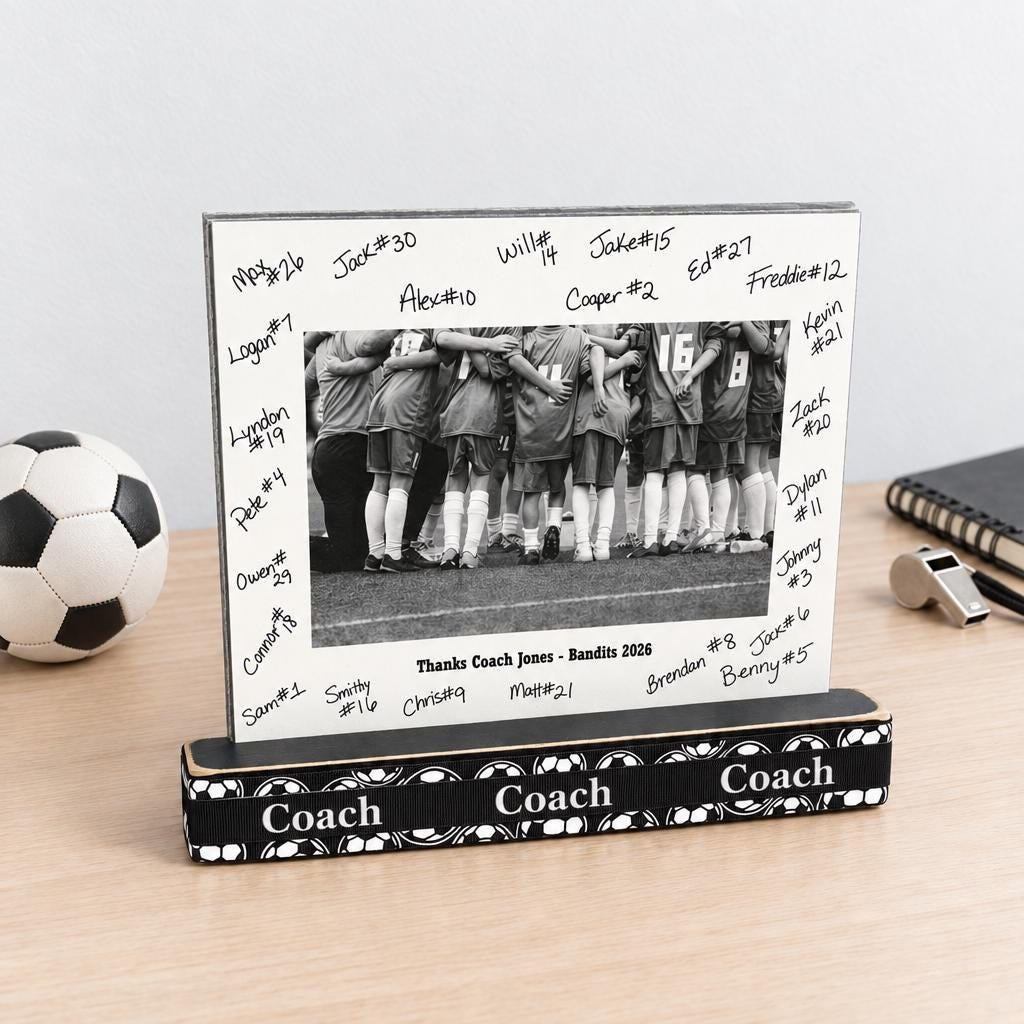 A soccer photo frame with a wooden base with soccer balls and COACH and a matted soccer photo with the team signatures all on the mat and custom team name and year as well.  This sits on a natural wooden desk along with a soccer ball, whistle and book.