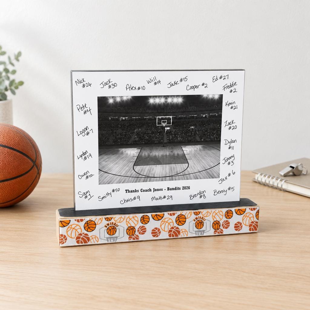 A basketball photo frame with a wooden base with basketballs and a matted basketball photo with the team signatures all on the mat and custom team name and year as well.  This sits on a natural wooden desk along with a basketball, whistle, book and a plant.
