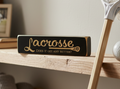 A rectangular black wooden sign with engraved lettering that reads 'Lacrosse Does it get any better?' resting on a light wood shelf, with a lacrosse stick and framed picture blurred in the background.