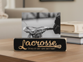 A black lacrosse-themed photo stand on a wooden table, holding a black-and-white photo of a lacrosse head. The sign reads 'Lacrosse' in engraved script with 'Does it get any better?' below, with books and a blurred indoor setting in the background.