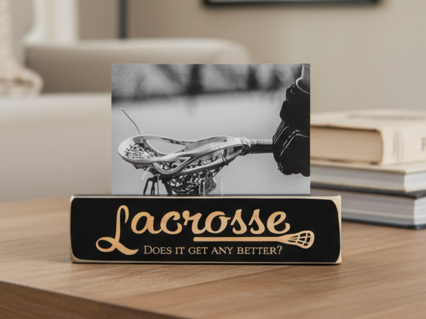 A black lacrosse-themed photo stand on a wooden table, holding a black-and-white photo of a lacrosse head. The sign reads 'Lacrosse' in engraved script with 'Does it get any better?' below, with books and a blurred indoor setting in the background.