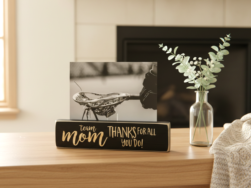 A decorative wooden sign on a light wood mantel with a black photo insert and lasered lettering that reads 'team mom THANKS FOR ALL YOU DO!'. A small clear glass vase with eucalyptus stands beside it, with a fireplace and neutral wall in the background.
