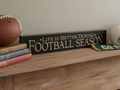 Black wooden sign with carved lettering "Life is better during FOOTBALL SEASON".  This is set on a table with other football decor.