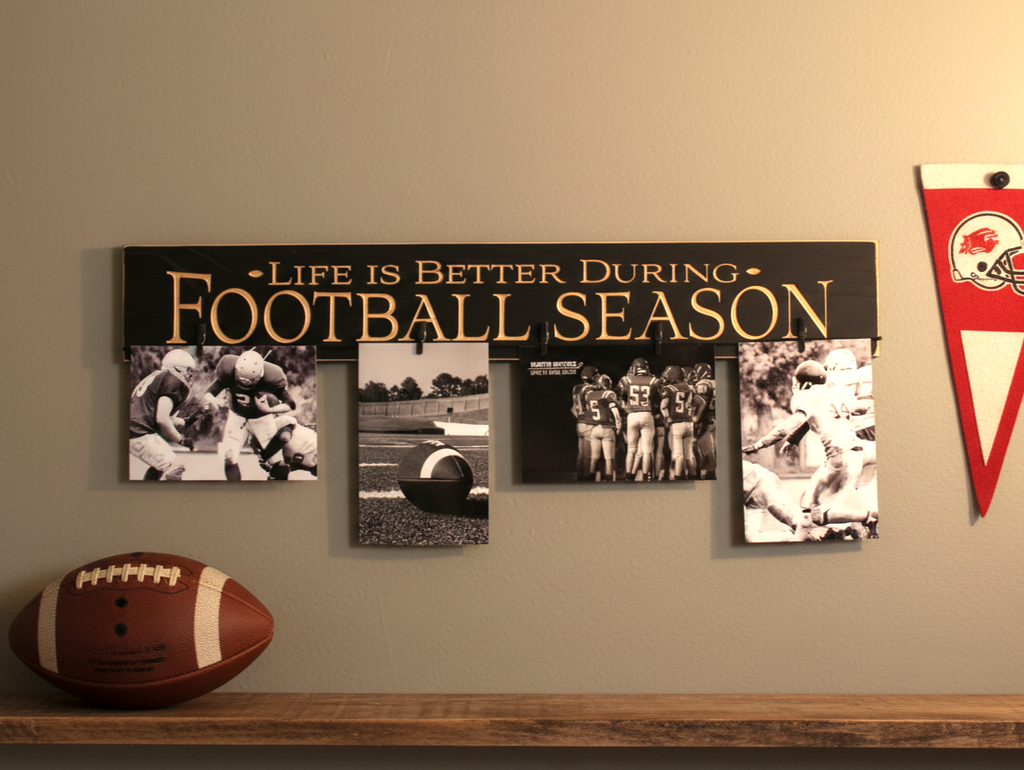 A wall-mounted football-themed photo collage sign with the text 'LIFE IS BETTER DURING FOOTBALL SEASON' in carved lettering on a black background, displayed above a wooden shelf with a football. The sign includes four black-and-white photo frames showing football players and a field.