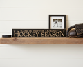 A rustic wooden shelf display featuring a black sign with engraved lettering that reads 'LIFE IS BETTER DURING HOCKEY SEASON,' a small black hockey puck, a framed black-and-white hockey photo, and a brown leather hockey glove against a white shiplap wall.