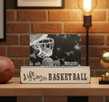 A rectangular wooden sign with a distressed finish that reads 'Life is better with BASKET BALL' in black lettering, styled on a wooden table next to a basketball and a gold trophy, with a black-and-white basketball photo behind it against a brick wall background.