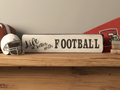 A rustic white wooden sign with black lettering that reads 'Life is better with FOOTBALL' displayed on a wooden shelf next to a football helmet, a football, and a red sports jersey.