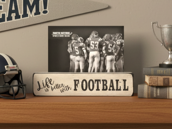 A decorative wooden sign on a shelf that reads 'Life is better with FOOTBALL' in black lettering, styled with a black-and-white football photo behind it, a helmet, books, and a trophy in a room setting.