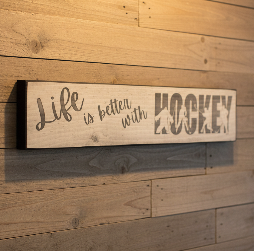 A rustic wooden sign with the phrase 'Life is better with HOCKEY' mounted on a wood-paneled wall. The left side reads 'Life is better with' in a cursive script, while the right side features the word 'HOCKEY' in a large block font filled with hockey players.