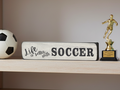 A rectangular white wooden sign with black lettering that reads 'Life is better with SOCCER' sits on a light wood shelf, flanked by a black-and-white soccer ball on the left and a gold soccer player trophy on the right.